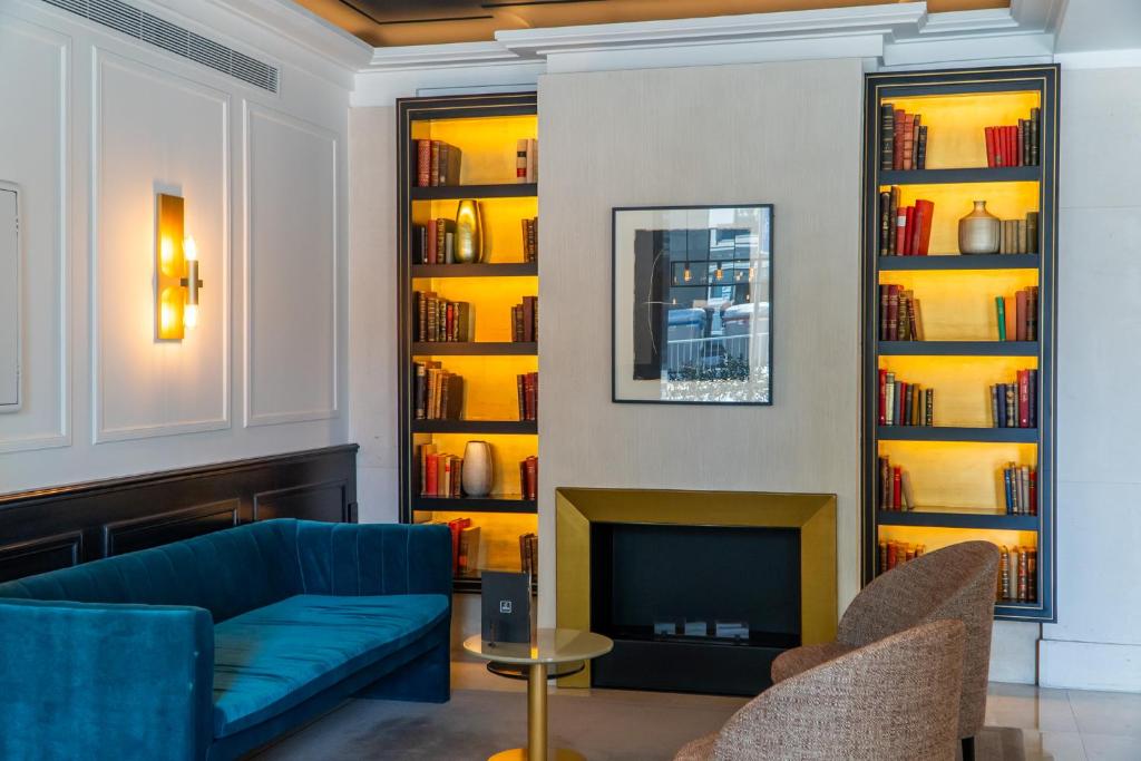 Mūsdienīgs viesnīcas interjers ar zilu dīvānu, divām krēslām un grāmatu plauktiem ar apgaismojumu. Siltas krāsas un eleganta dizaina elementi rada patīkamu atmosfēru.