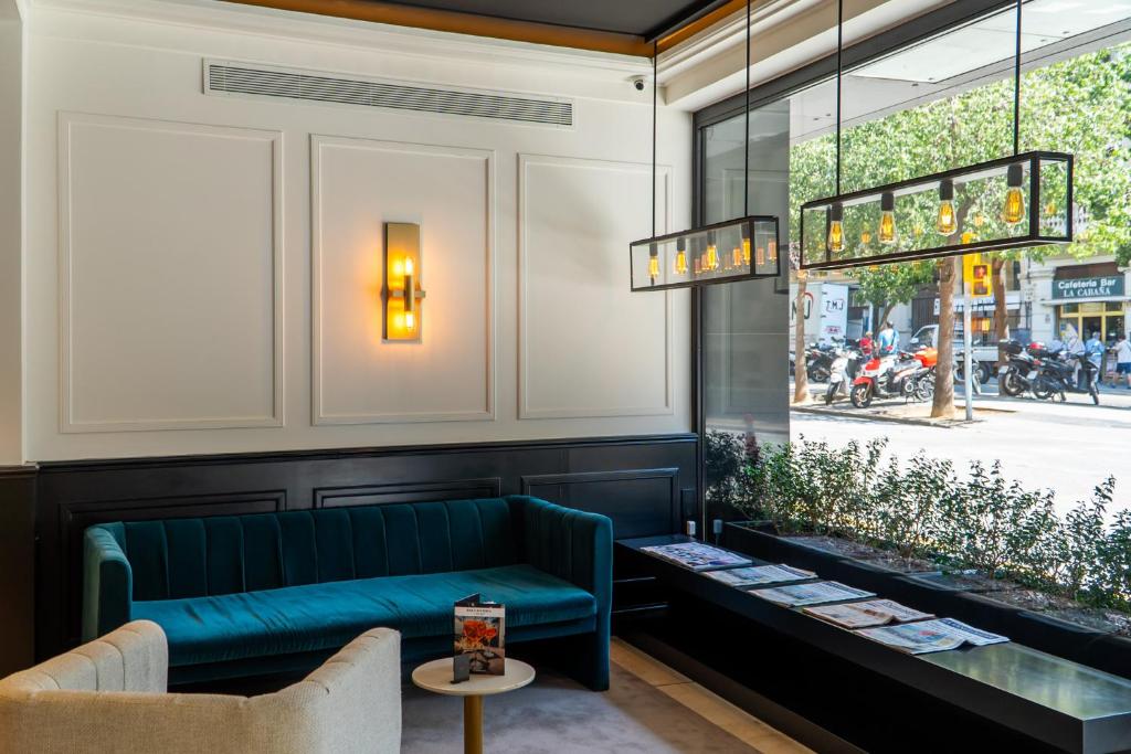 Elegants viesistabas interjers ar zaļu dīvānu un modernu apgaismojumu, kas sniedzas līdz plašajiem logiem, caur kuriem redzama iela ar motocikliem un kokiem.