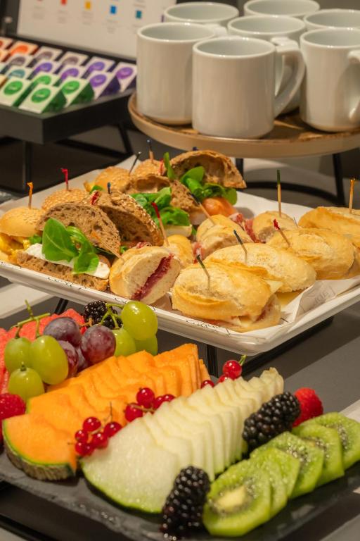 Svaigs uzkodu galds ar dažādām maizēm, augļiem un dzērieniem, piemērots sanāksmēm un pasākumiem. Uzkodas ietver veselīgas iespējas, piemēram, augļus un maizītes ar dažādām pildījumiem.