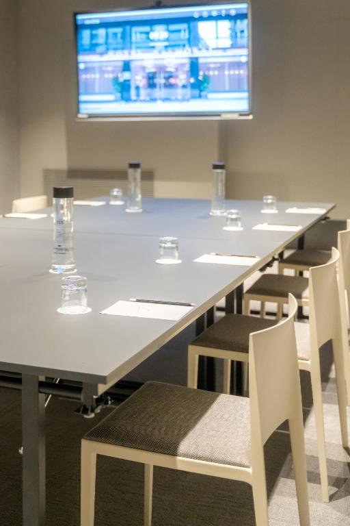 Moderns sapulču zāles interjers ar garu galdu, krēsliem un ekrānu prezentācijām. Uz galda ir ūdens pudeles un piezīmju lapas.