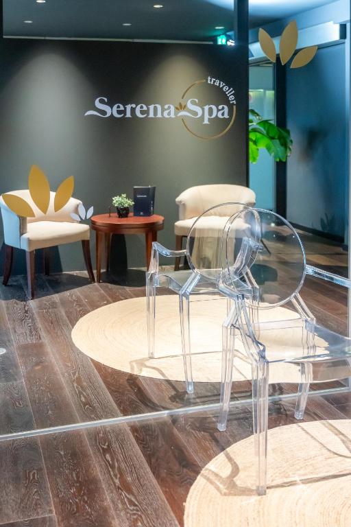 Mūsdienīgs spa interjers ar caurspīdīgu krēslu un ērtām mēbelēm, radot relaksējošu atmosfēru. Uz sienas redzams "Serena Spa" logo.