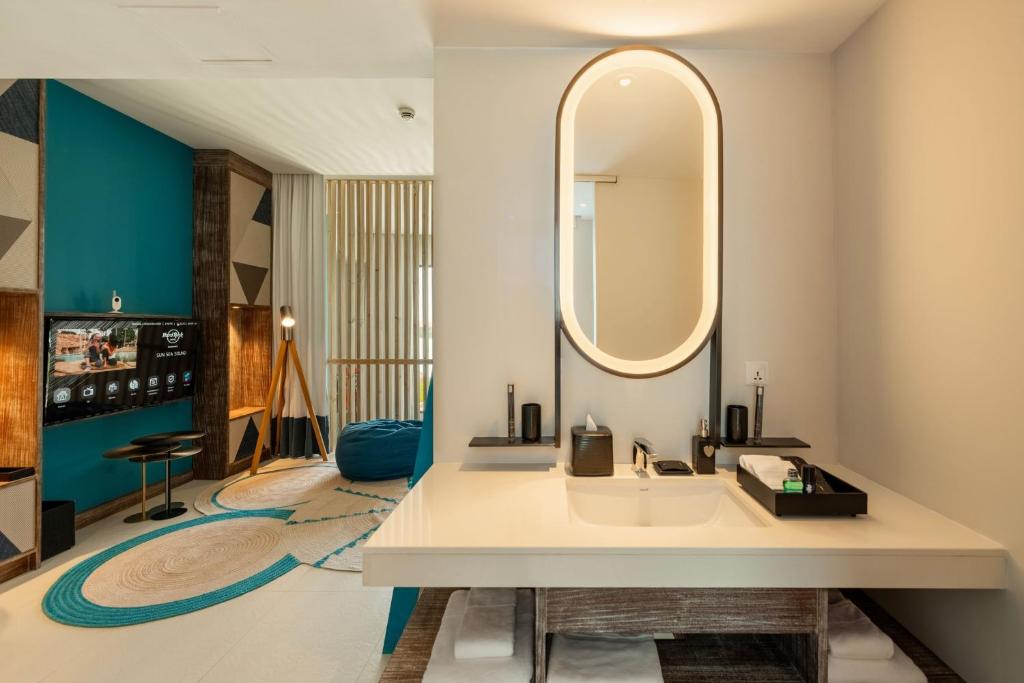 Mūsdienīgs vannas istabas interjers ar elegantu izlietni un apaļu spoguli, papildināts ar krāsainu dzīvojamo telpu un modernu televizoru.