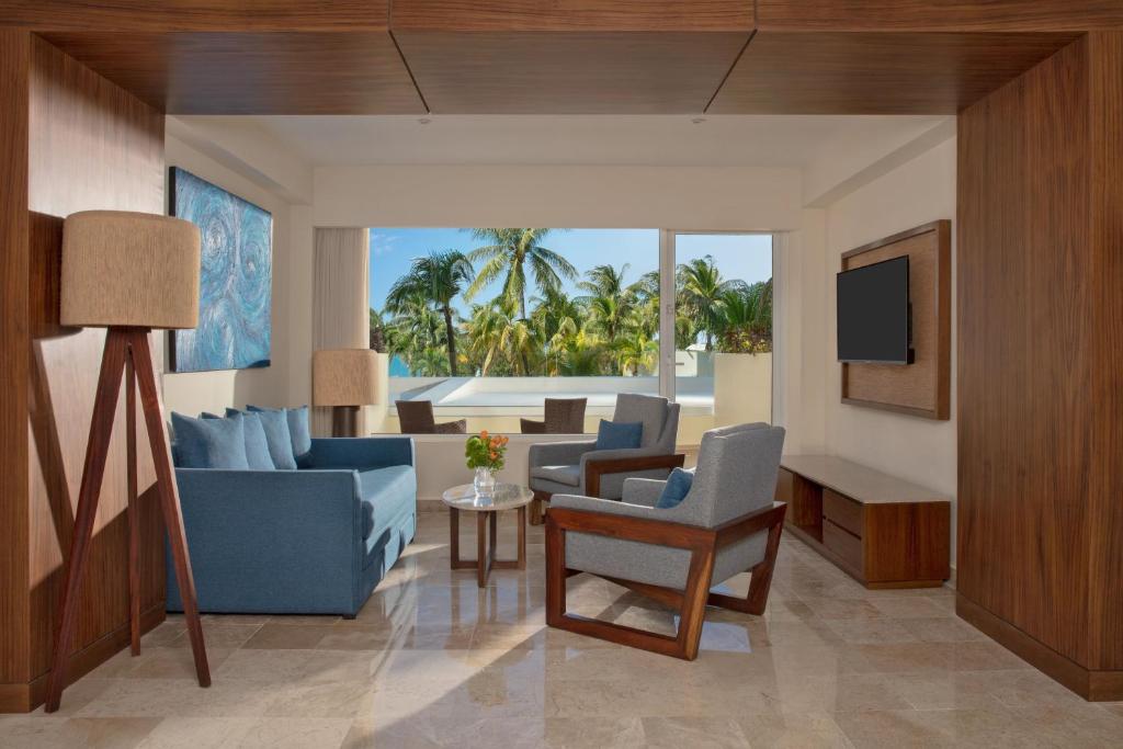 Mūsdienīgs viesnīcas numurs ar skatu uz palmu dārzu, kurā ir ērti dīvāni un gaismas objekts. Interjers ir veidots ar siltām koka un gaišām krāsām.