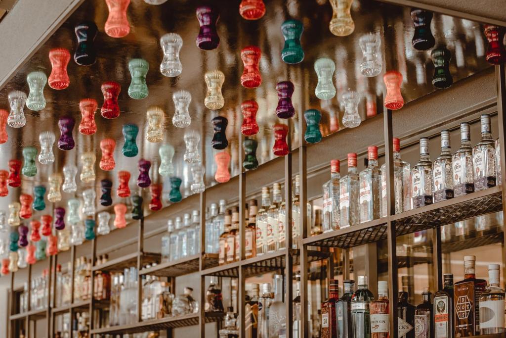 Krāsains bāra interjers ar gaismas elementiem, kas atrodas pie griestiem, un dažādām alkoholisko dzērienu pudelēm uz plauktiem.