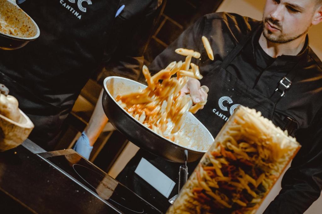 Pavārs, tērpies melnā, sajauc makaronus ar mērci lielā pannā, kamēr otra persona stāv blakus. Fons ir tumšs, ar pasta trauku tuvplānā.