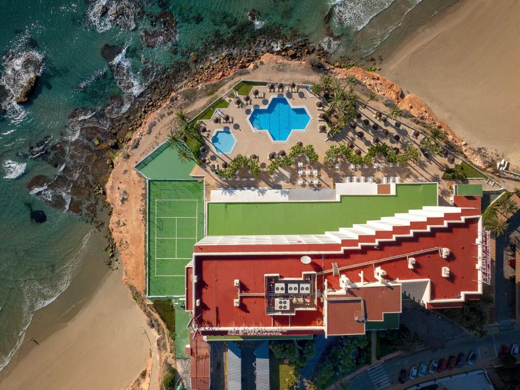 Lidojums virs viesnīcas ar baseinu un tenisa laukumiem, kas atrodas netālu no pludmales un krasta līnijas. Apkārtnē redzamas palmas un akmeņainā jūras mala.