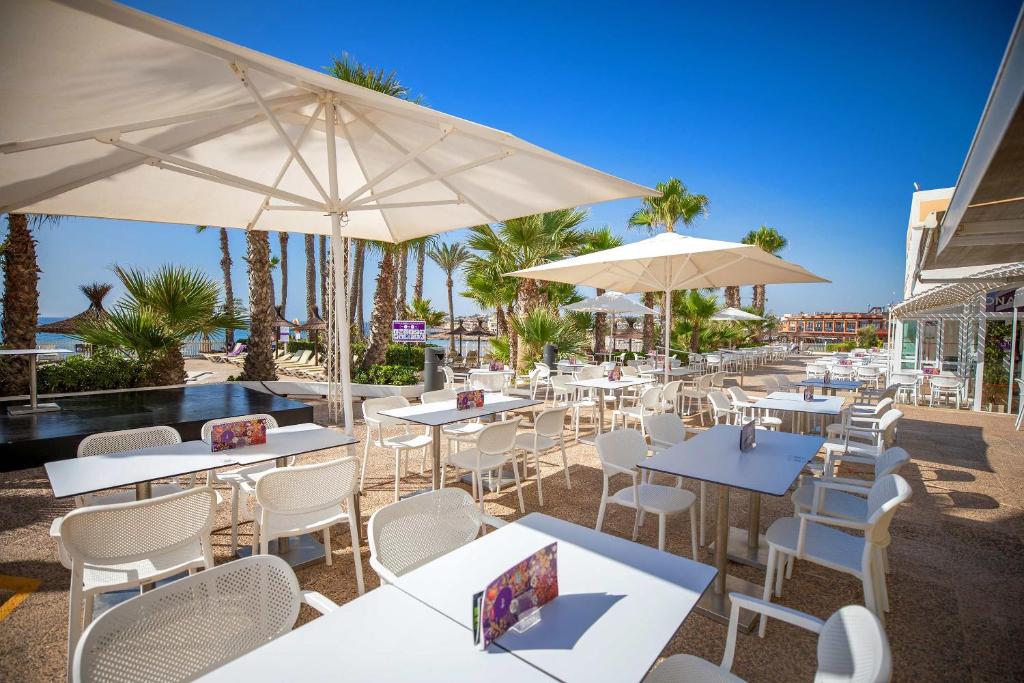 Atvērtā terase ar baltām galdiem un krēsliem, apstādīta ar palmām, un skats uz pludmali. Saulaina diena ar skaidru debesu fona.