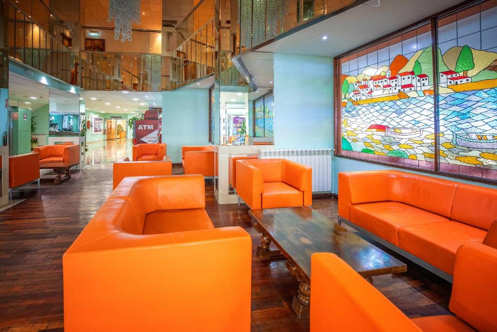 Mūsdienīgs un krāsains viesnīcas interjers ar oranžiem dīvāniem un koka galdu, kas rada viesmīlīgu atmosfēru. Logu vitrāža attēlo gleznainu ainavu ar mājām un laivām pie ūdens.