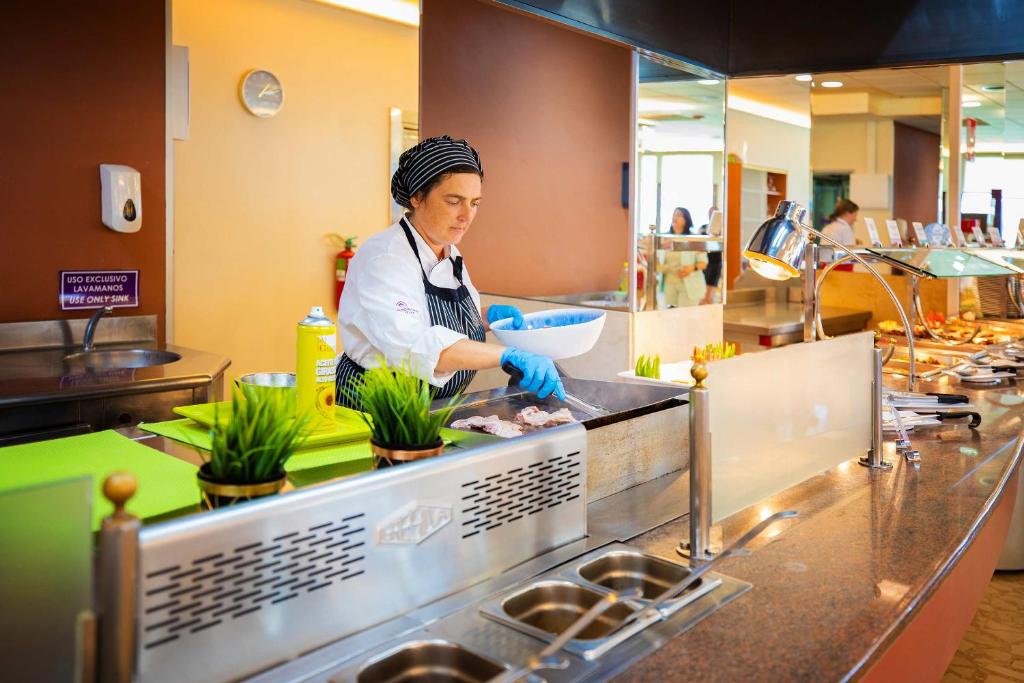 Konditorejas darbiniece, tērpusies balta priekšautā un cepurē, gatavo ēdienu restorānā, izmantojot profesionālo virtuves aprīkojumu. Fons satur dažādas ēdienu izvēles un cilvēkus, kas apmeklē bufeti.