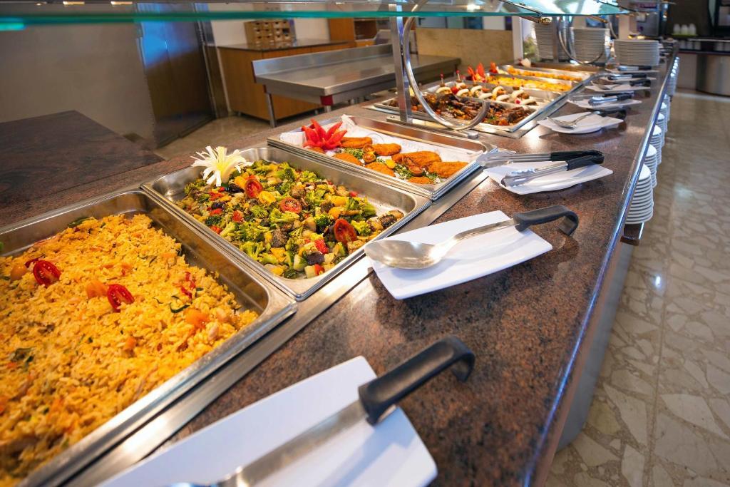 Bagātīgs bufetes galds ar dažādiem ēdieniem, tostarp dārzeņu salātiem, rīsiem un ceptiem ēdieniem. Galds ir sakārtots ar traukiem un piederumiem, radot viesmīlīgu atmosfēru.