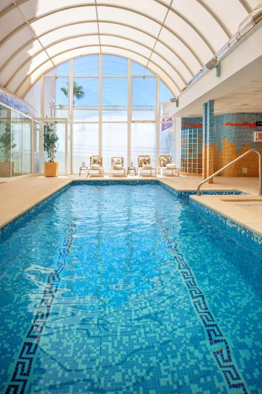 Iekštelpu peldbaseins ar caurspīdīgu kupolu un saulainu gaismu, apkārt peldbaseinam ir ērtas atpūtas krēsli.