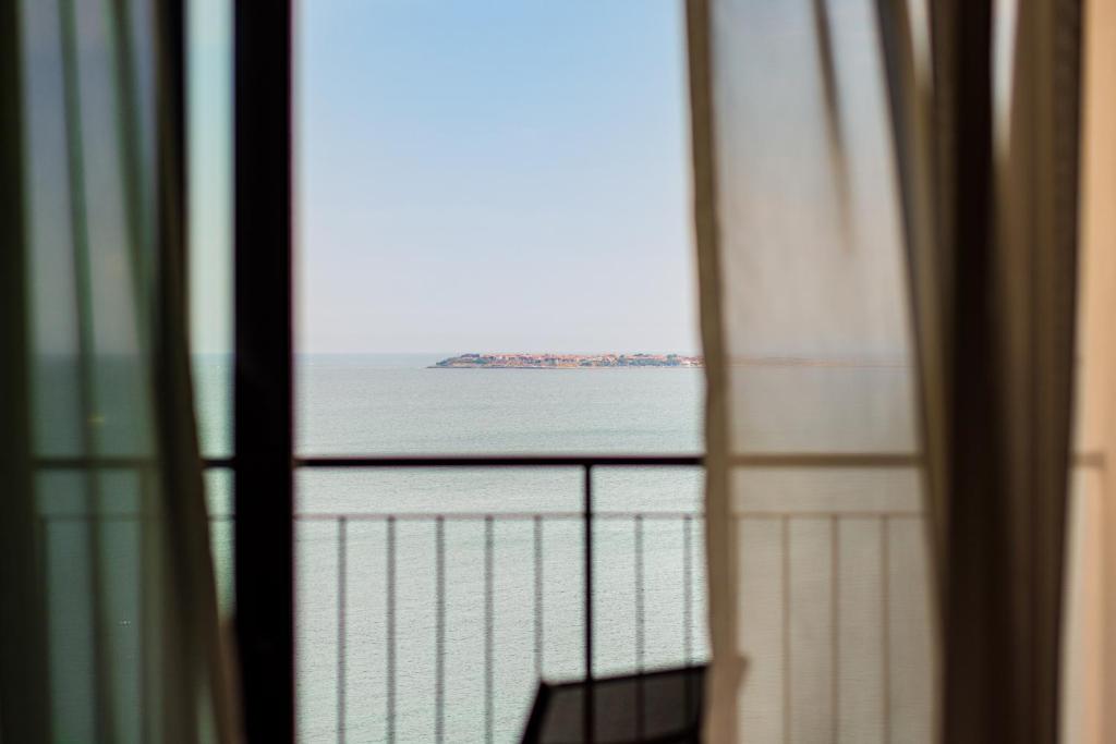 Skats uz jūru caur aizvērtām žalūzijām, redzama maza sala tālumā.