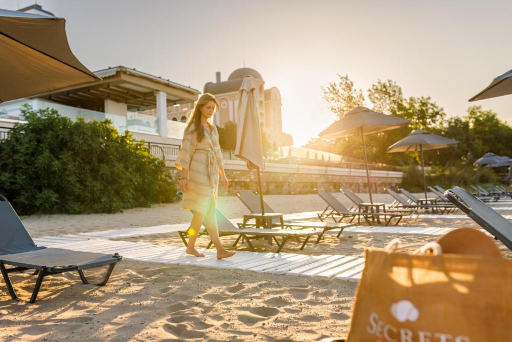 Jauna sieviete pastaigājas pa pludmali saulrietā, apņemta ar siltām gaismām un pludmales krēsliem. Attēlā redzama arī pludmales soma ar "Secrets" logo.