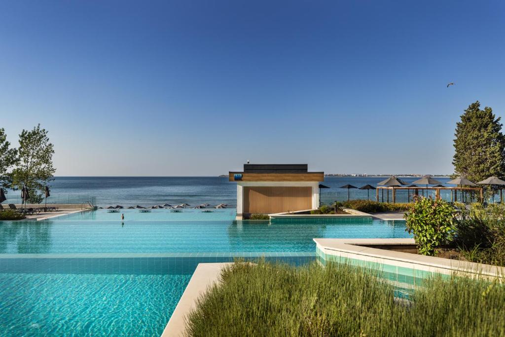 Lielisks skats uz modernu peldbaseinu, kas atrodas pie jūras, ar ziliem ūdeņiem un skaidru debesu fonu. Apkārt peldbaseinam ir augi un sauļošanās vietas ar saules sargiem.