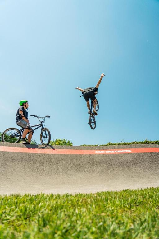 Divi jaunieši izpilda BMX trikus saulainā dienā, viens no viņiem lido gaisā, bet otrs novēro no velosipēda.