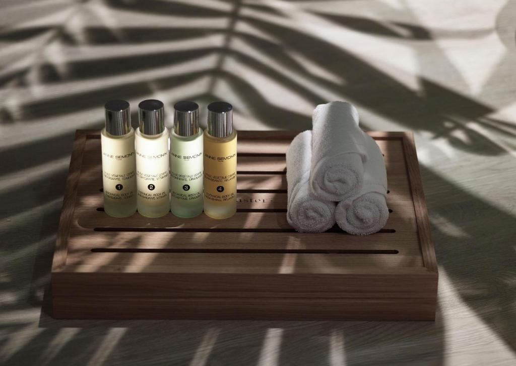 Uz koka paletes novietoti četri aromātiskie eļļu flakoni un trīs sarullēti dvieļi, izceļot relaksējošu atmosfēru. Gaisā redzamas palmu ēnas, kas piešķir attēlam eksotisku noskaņu.