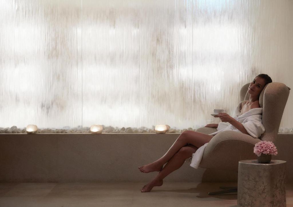 Sieviete relaksējas modernā spa telpā, tērpta baltā dvielī, turot krūzi ar dzērienu. Fons ir maigs ar ūdens plūsmu un akmeņiem, radot mierīgu atmosfēru.