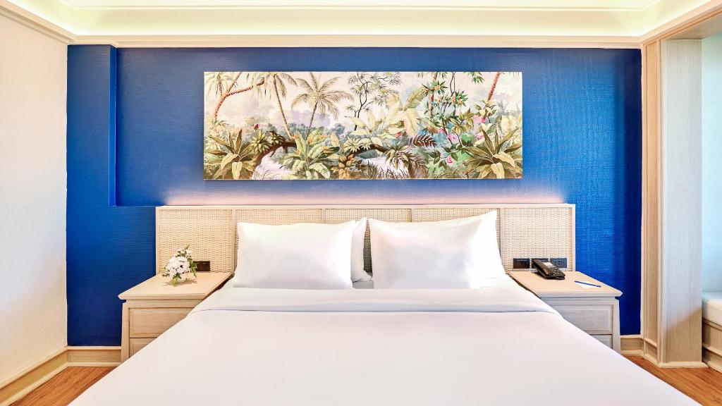 Mūsdienīga guļamistaba ar zilu sienu un ainavu gleznu virs gultas, kurā ir balti spilveni un gultas veļa. Sānos redzama koka naktsgaldiņš ar dekoratīvu ziedu.