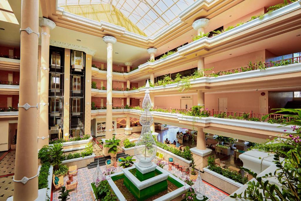 Moderns viesnīcas vestibils ar elegantām kolonnām, augstiem griestiem un plašu augstāku stāvu terasi, kurā redzama zaļā apstādījumu un strūklakas kombinācija.