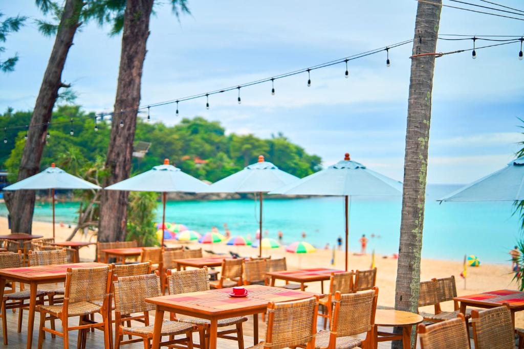 Pludmales restorāns ar koka galdiem un krēsliem, kas atrodas zem lieliem lietussargiem, ar skatu uz zilo jūru un smiltīm. Apkārt redzami pludmales apmeklētāji un krāsaini lietussargi.