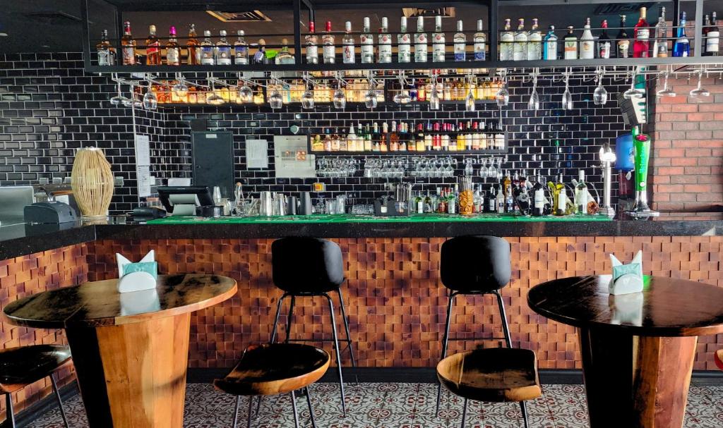 Mūsdienīgs bārs ar elegantu interjeru, kurā redzami koka galdi un melnā keramikas flīžu siena. Bārā ir plašs alkohola klāsts un glāzes, kas izkārtotas uz plauktiem.