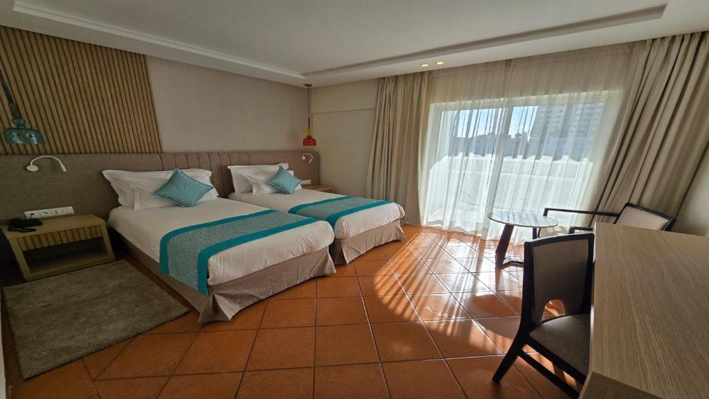 Mūsdienīgs viesnīcas numurs ar divām gultām, dekorēts ar zilām spilveniem un koka mēbelēm, plašs logs ar aizkariem un skatu uz ārpusi.