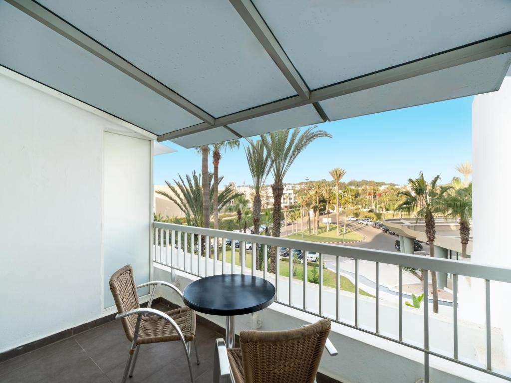 Balkons ar skatu uz zaļo ainavu, kur redzami palmas un ceļi. Pie galda ir divas krēslu vietas, radot relaksējošu atmosfēru.