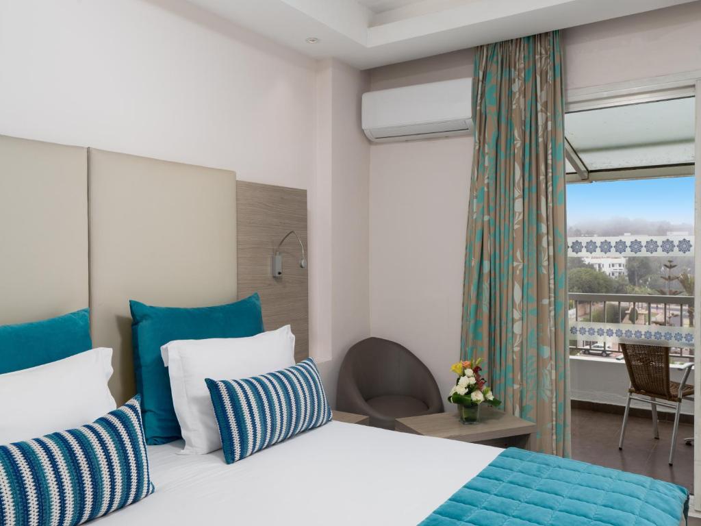 Mūsdienīga viesnīcas numura interjers ar gaišām krāsām, divām gultām ar spilveniem un skatu uz balkonu. Numurā ir ērtas mēbeles un dekoratīvi akcenti.