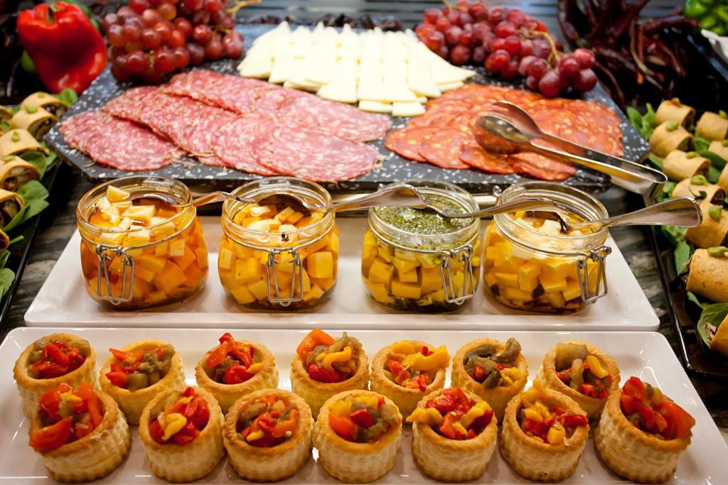 Svinību galda izkārtojums ar dažādiem uzkodu elementiem, tostarp desām, sieriem, marinētiem dārzeņiem un mini kārtainajām mīklām. Galvenajā plāksnē ir krāsaini augļi un dārzeņi, kas papildina piedāvājumu.