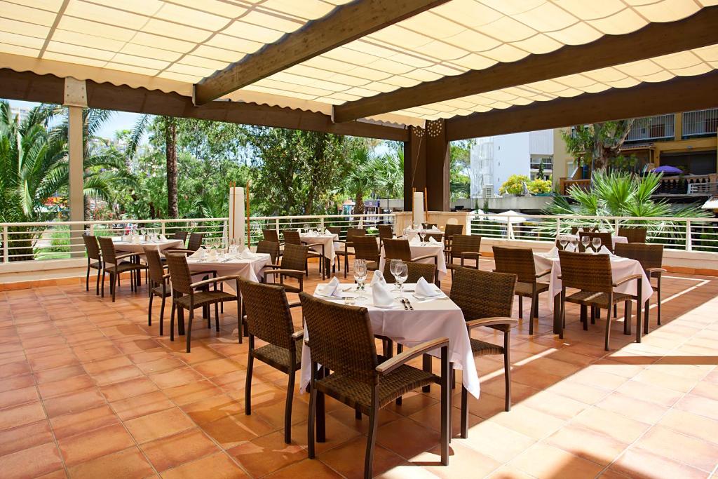 Āra restorāna terase ar elegantām galdiem un krēsliem, kas izvietoti zem saulessarga. Apkārt redzami zaļumi un palmām, radot relaksētu gaisotni.