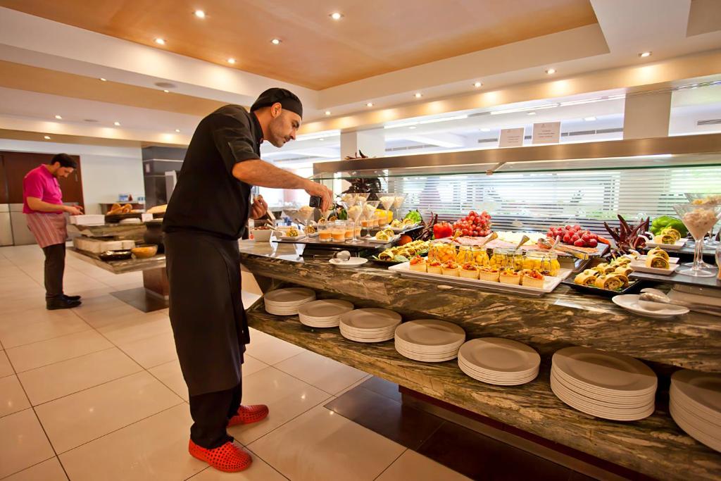 Bufete ar plašu ēdienu klāstu, kurā redzams viesmīlis, kas pasniedz ēdienus, un otra persona, kas gatavo. Galda virsma ir piepildīta ar augļiem un desertiem.