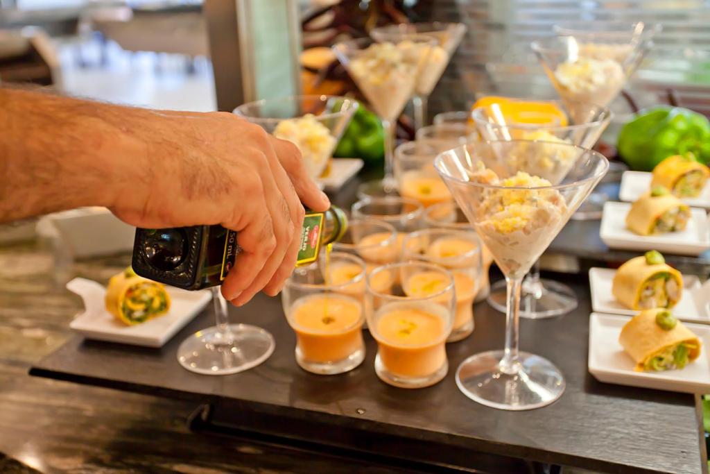 Gatavošanās viesību uzkodām, kurās redzami dažādi dzērieni un uzkodas, kam pievieno olīveļļu.