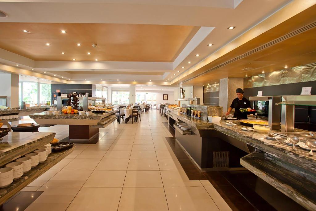 Moderns bufetes restorāns ar plašu ēdienu izvēli, apgaismotu telpu un sēdvietām viesiem. Redzams šefpavārs, kas gatavo ēdienus.