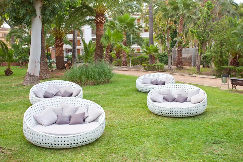 Āra mēbeles ar baltām apaļām sēdvietām un spilveniem, novietotas zaļajā zālē, apstādījumos ar palmām un citiem augiem.