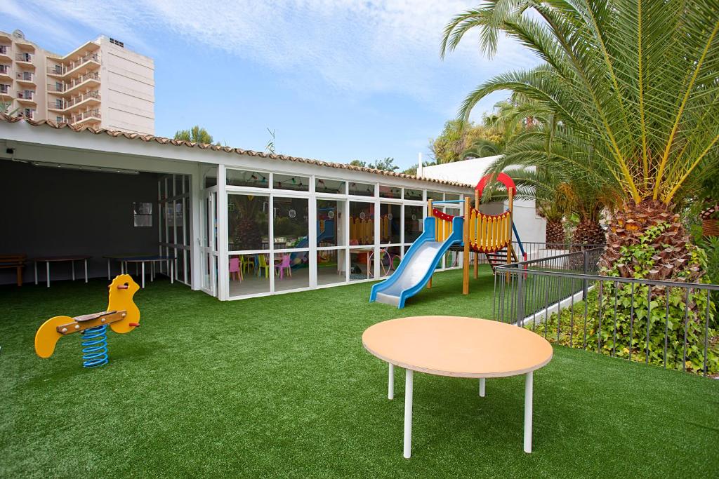 Bērnu rotaļu laukums ar šūpolēm un slidkalniņu, zaļā zālīte un krāsains galdiņš. Apkārt aug palm koku.