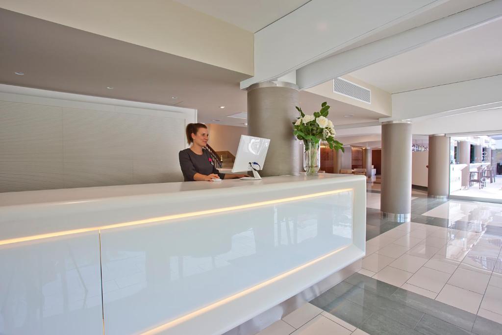 Mūsdienīgs viesnīcas reģistratūras galds ar laipnu darbinieku, kas apkalpo viesus. Interjerā ir gaišas krāsas un eleganti ziedi.