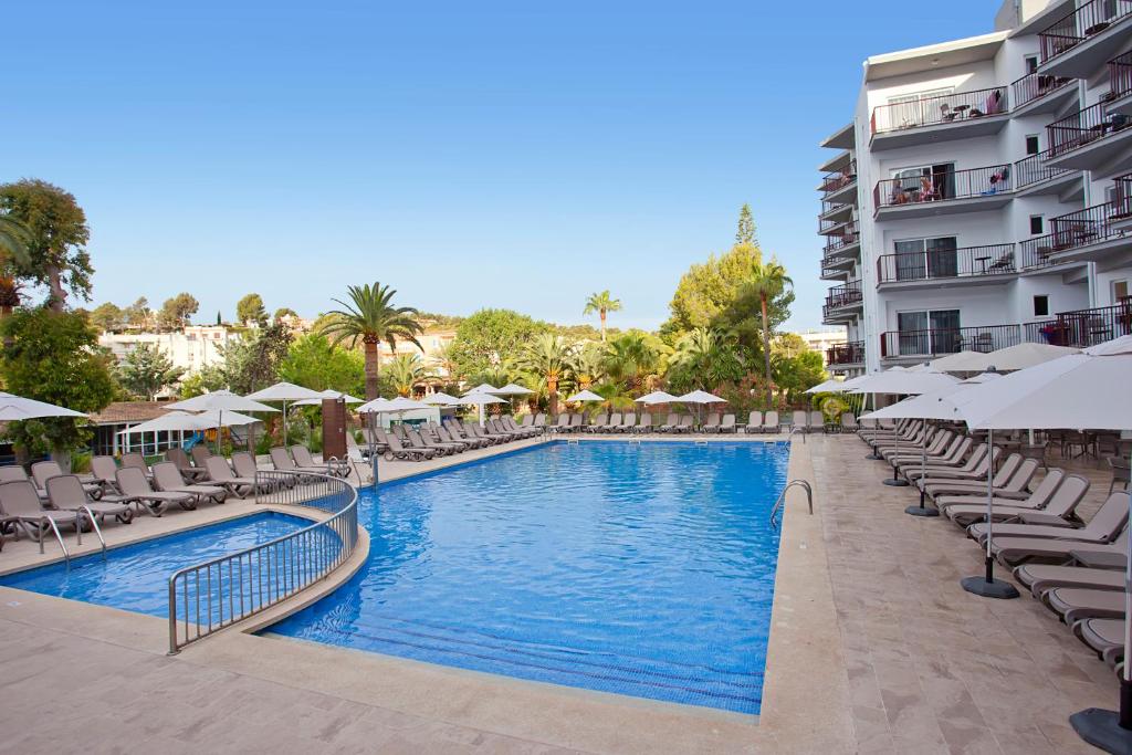 Viesnīcas baseins ar sauļošanās krēsliem un saulessargiem, apkārtējiem palmu kokiem un zaļo dārzu. Ideāla vieta atpūtai un relaksācijai saulainā dienā.