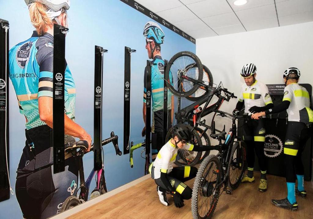 Velosipēdisti strādā pie velosipēdu apkopes telpā ar modernu dizainu un uz sienas attēlotām sporta ainavām.