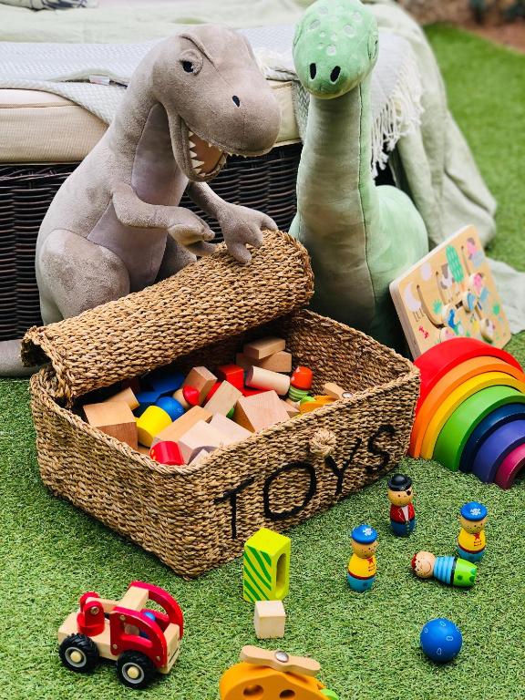 Rotaļlietu kaste, kas pilna ar krāsainām koka rotaļlietām, apņemta ar mīkstiem dinozauriem, veido rotaļīgu un krāsainu bērnu spēļu vidi.