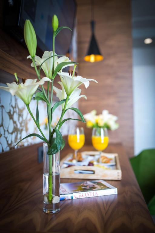 Svaigas baltas lilijas glāzē, uz koka galda, blakus oranžām dzēriena glāzēm un žurnālu "Kroātija" ar uzkodu šķīvi.