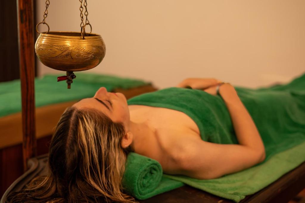 Masažas ar tradicionālo eļļu, sieviete atpūšas uz galda ar zaļu dvieli, virs viņas karājas zelta trauks.