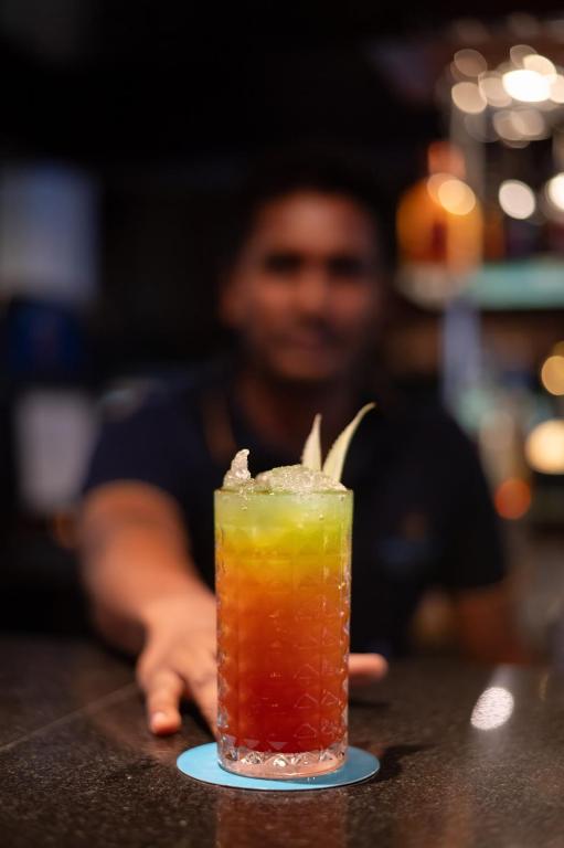 Krāsains kokteilis ar zaļas un sarkanas nokrāsas, rotāts ar zaļu augu, novietots uz bāra. Fons izplūdis, uzsverot dzēriena pievilcību.