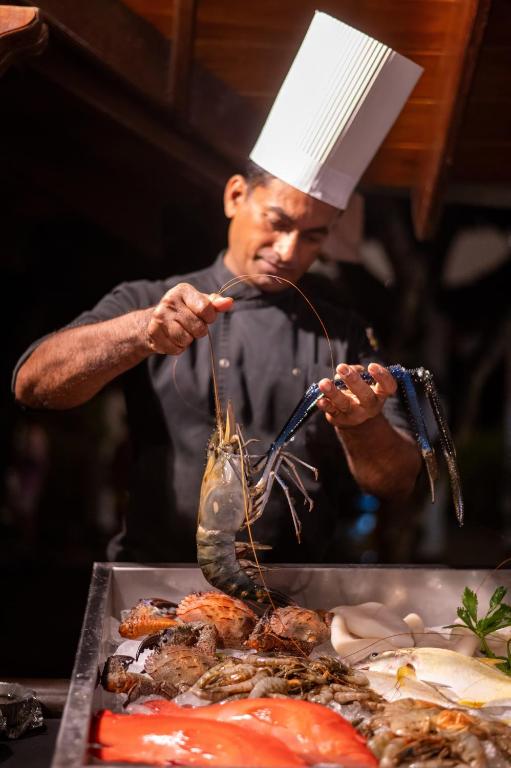 Šefpavārs gatavo jūras veltes, paceldams lielu garneli, kamēr ap viņu ir dažādi jūras produkti.