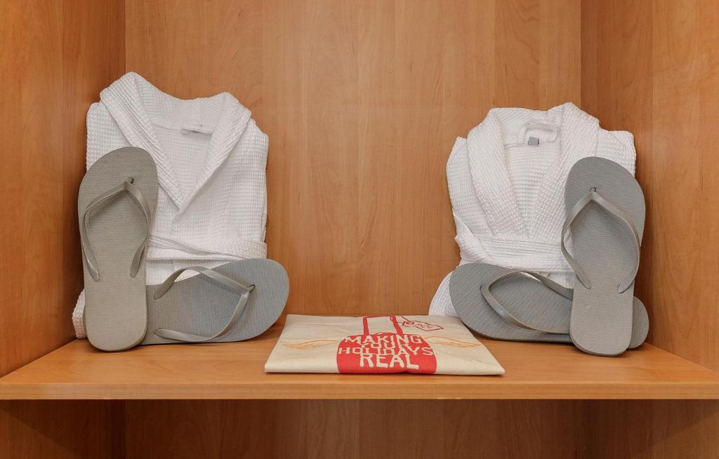 Baltas peldmētelis un pelēki flip-flops, sakārtoti uz koka plaukta, blakus ir soma ar uzrakstu "MAKING HOLIDAYS REAL".