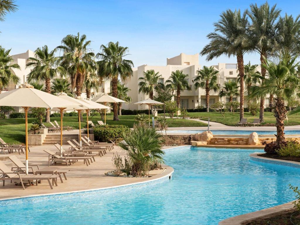 Peldbaseins ar palmu kokiem un sauļošanās krēsliem, radot relaksējošu atpūtas vidi. Labi apzaļumots apvidus ar saulessargiem un viesnīcas ēkām fona.