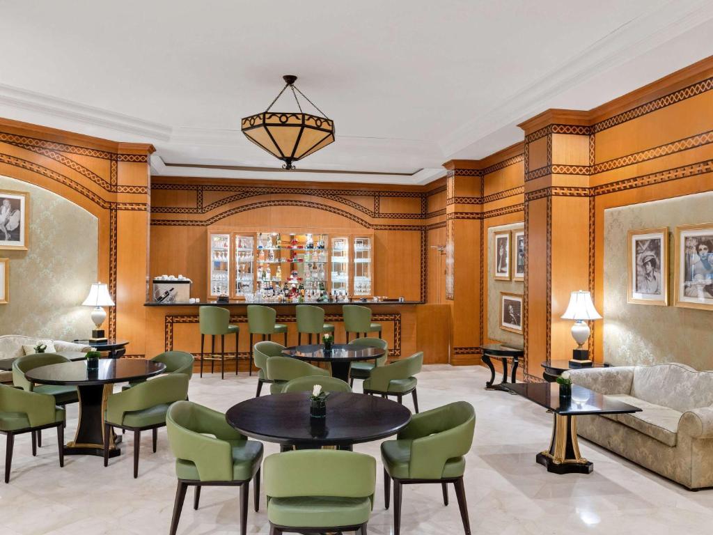 Elegants bārs ar koka apdari un mājīgu sēdvietu zonu, kurā ir zaļi krēsli un melni galdi. Aizmugurē ir bāra plaukts ar dzērieniem un gaismas lampas, kas piešķir siltumu telpai.