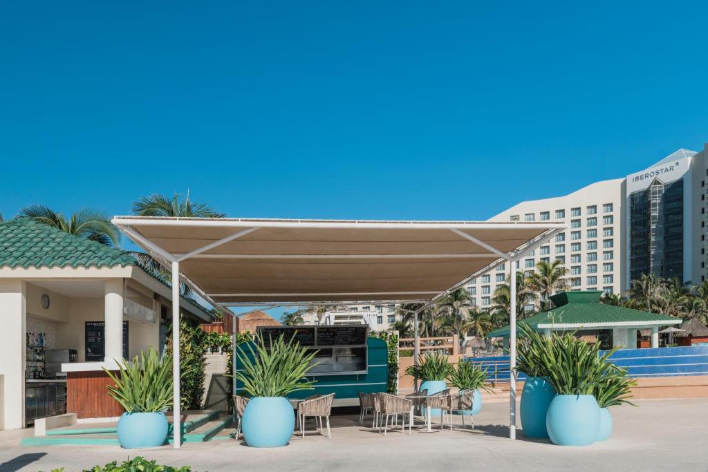 Tropu atpūtas zona ar bāru un sēdvietām, ieskauta zilo puķu podos un palmām, pret debesu zilu fonu.