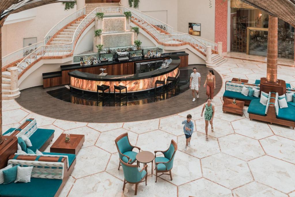 Mūsdienīgs viesnīcas vestibilis ar elegantu bāru, ap kuru staigā ģimene. Interjerā ir spožs marmora grīdas segums un stilīgas mēbeles, kas rada relaksējošu atmosfēru.