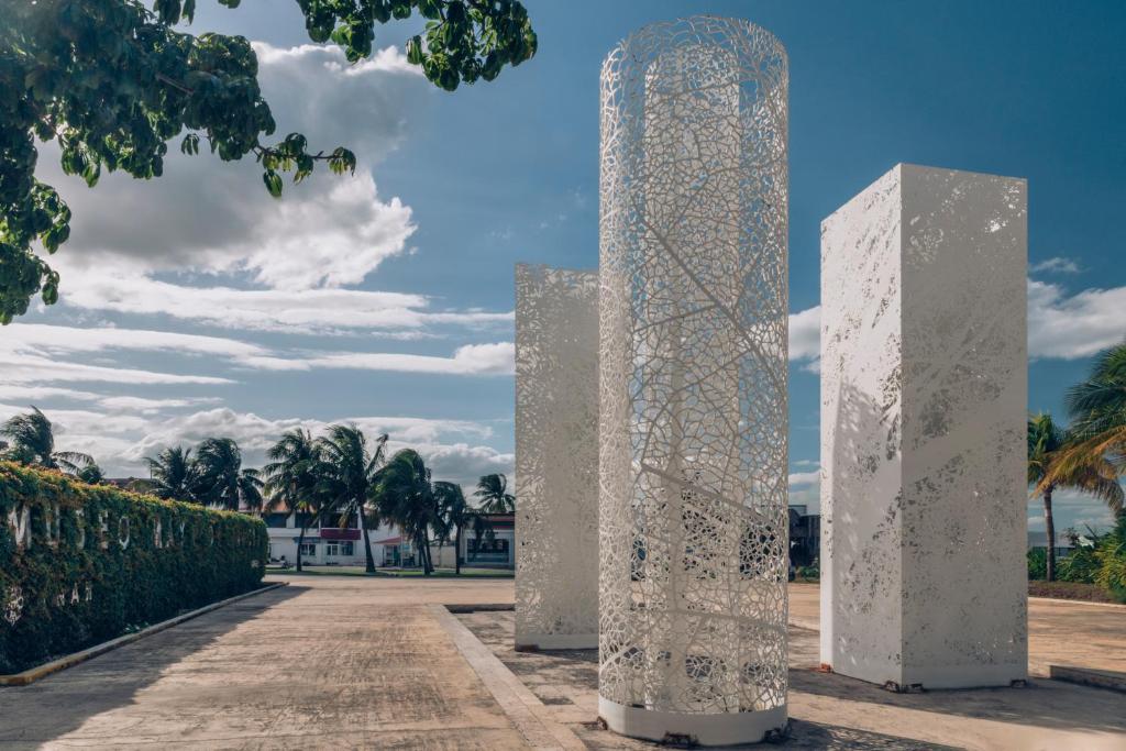 Mūsdienīga arhitektūras instalācija ar baltiem stabiem, kas izgatavoti no perforēta metāla, un skaista dabas ainava ar palmām un zilo debesu fonu.