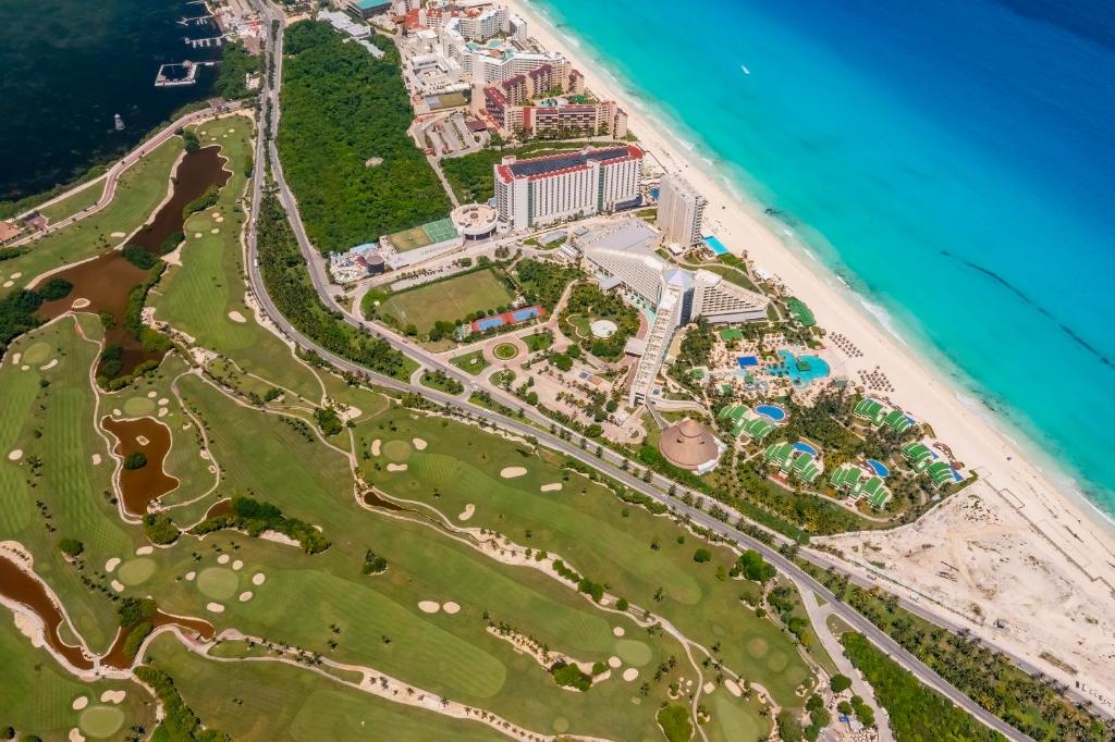 Gaisa skats uz pludmali un kūrortiem, kas atrodas netālu no zaļām golfa laukumiem un zilajiem ūdeņiem. Šī ainava apvieno dabu un luksusa atpūtu, piedāvājot lielisku atpūtas vietu.